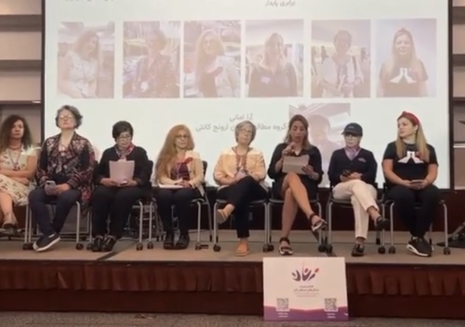 Eight women speakers seated on a conference stage in front of a screen, one speaking into a microphone